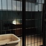 Interior de uma cela prisional com grades, pia e mesa em iluminação baixa.