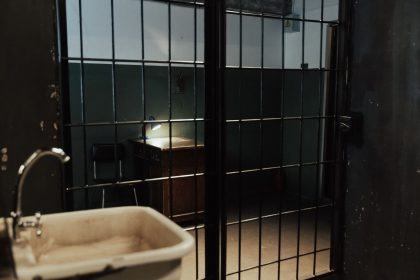 Interior de uma cela prisional com grades, pia e mesa em iluminação baixa.