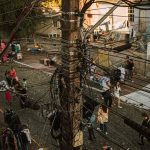 Rua movimentada com emaranhado de cabos de telecomunicação sobre as edificações e pessoas circulando no local.
