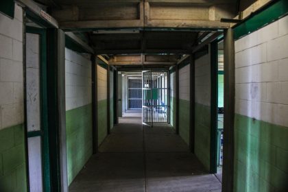 Corredor vazio de uma prisão, com portas de celas abertas e grades metálicas.