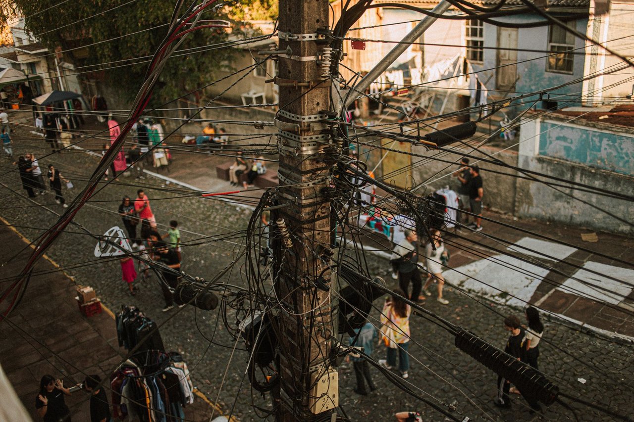Rua movimentada com emaranhado de cabos de telecomunicação sobre as edificações e pessoas circulando no local.
