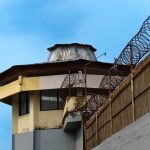 Torre de vigilância de uma unidade prisional cercada por arame farpado sob céu azul.