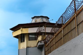 Torre de vigilância de uma unidade prisional cercada por arame farpado sob céu azul.